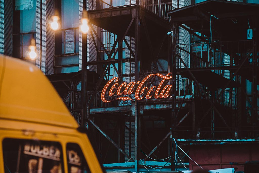 Dramatic urban street scene featuring a neon Coca Cola sign on an industrial building.